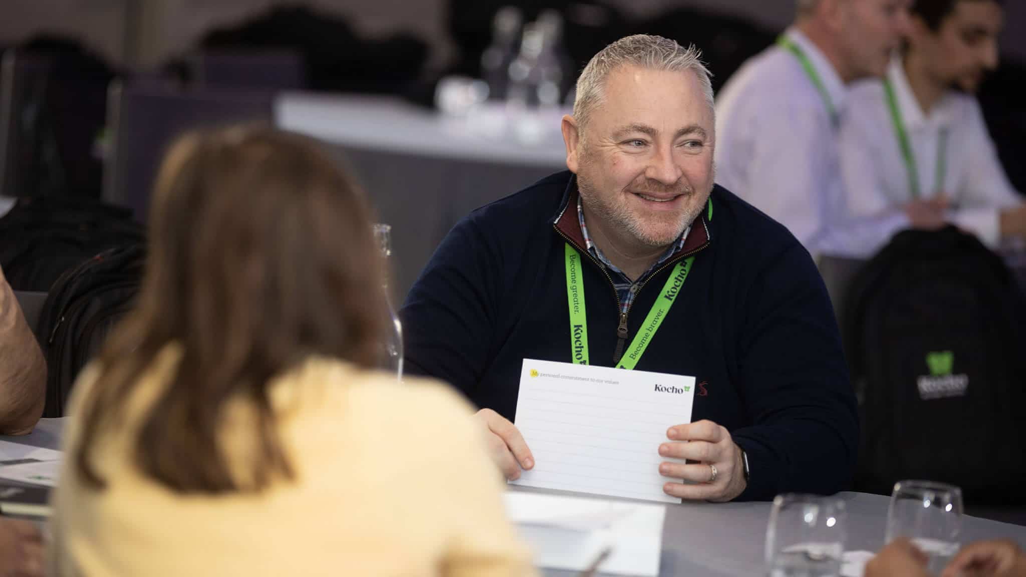 Man with grey hair sat at table smiling and holding white card