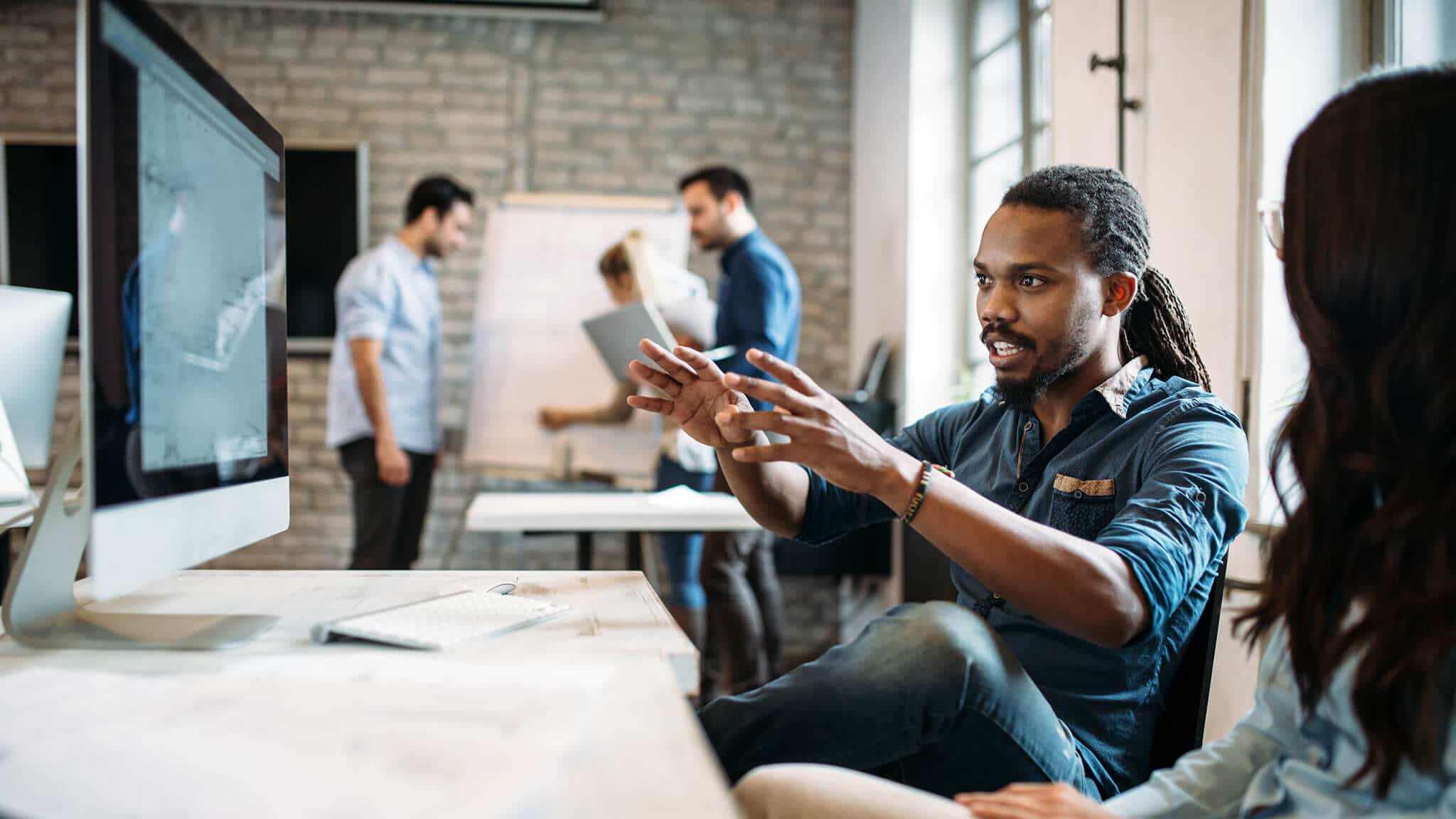 Man in blue shirt with dreadlocks gesturing towards desktop screen