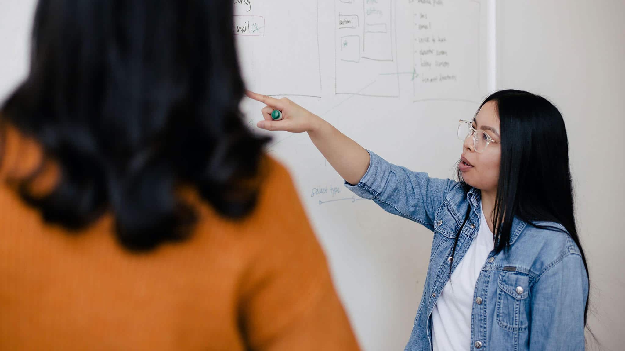 Woman wearing glasses with black hair in denim jacket pointing at whiteboard